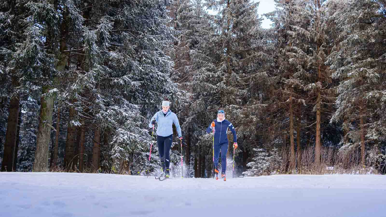 Twee personen doen aan langlaufen door een besneeuwd landschap. Ze dragen warme functionele kleding, wintermutsen en gebruiken skistokken. De omgeving is gekenmerkt door met sneeuw bedekte bomen en een heldere winterse sfeer.