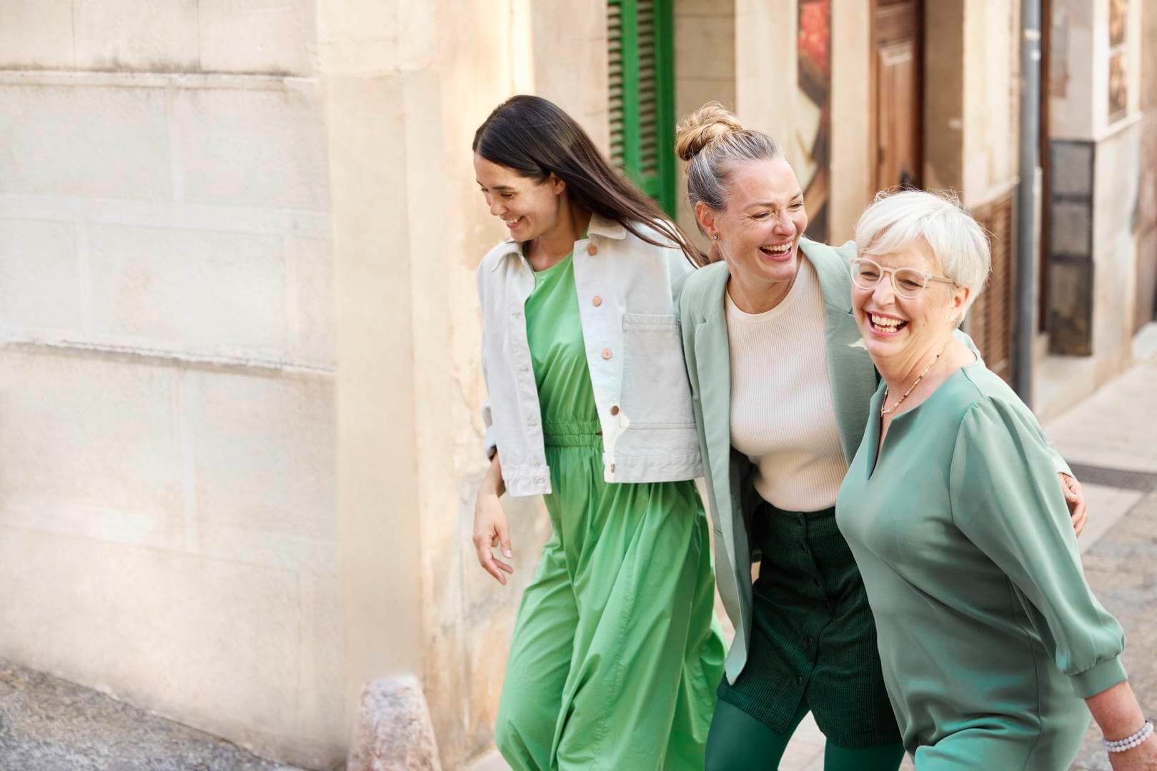 Drie vrouwen van verschillende generaties lachen samen, terwijl ze arm in arm door een mediterrane oude binnenstad lopen. Ze dragen stijlvolle outfits in verschillende tinten groen die harmonieus op elkaar zijn afgestemd.