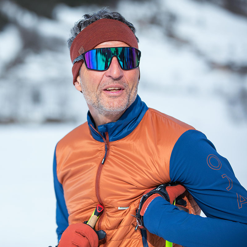 Een oudere man met een skibril, een bruine hoofdband en een oranje-blauwe winterjas werd gefotografeerd in een portret voor een wit bos.