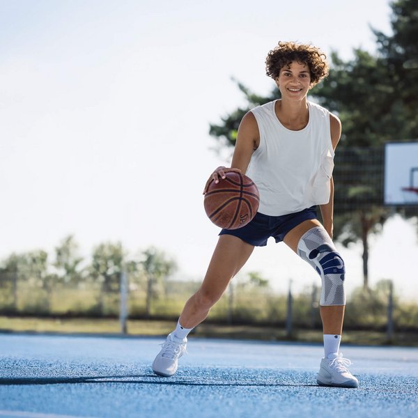 Jonge vrouw met wit hemdje, donkerblauwe sportbroek en witte kniebandage speelt basketbal op een buitenveld met blauwe ondergrond; ze dribbelt de bal en lacht naar de camera, op de achtergrond zijn een basketbalring en bomen te zien.
