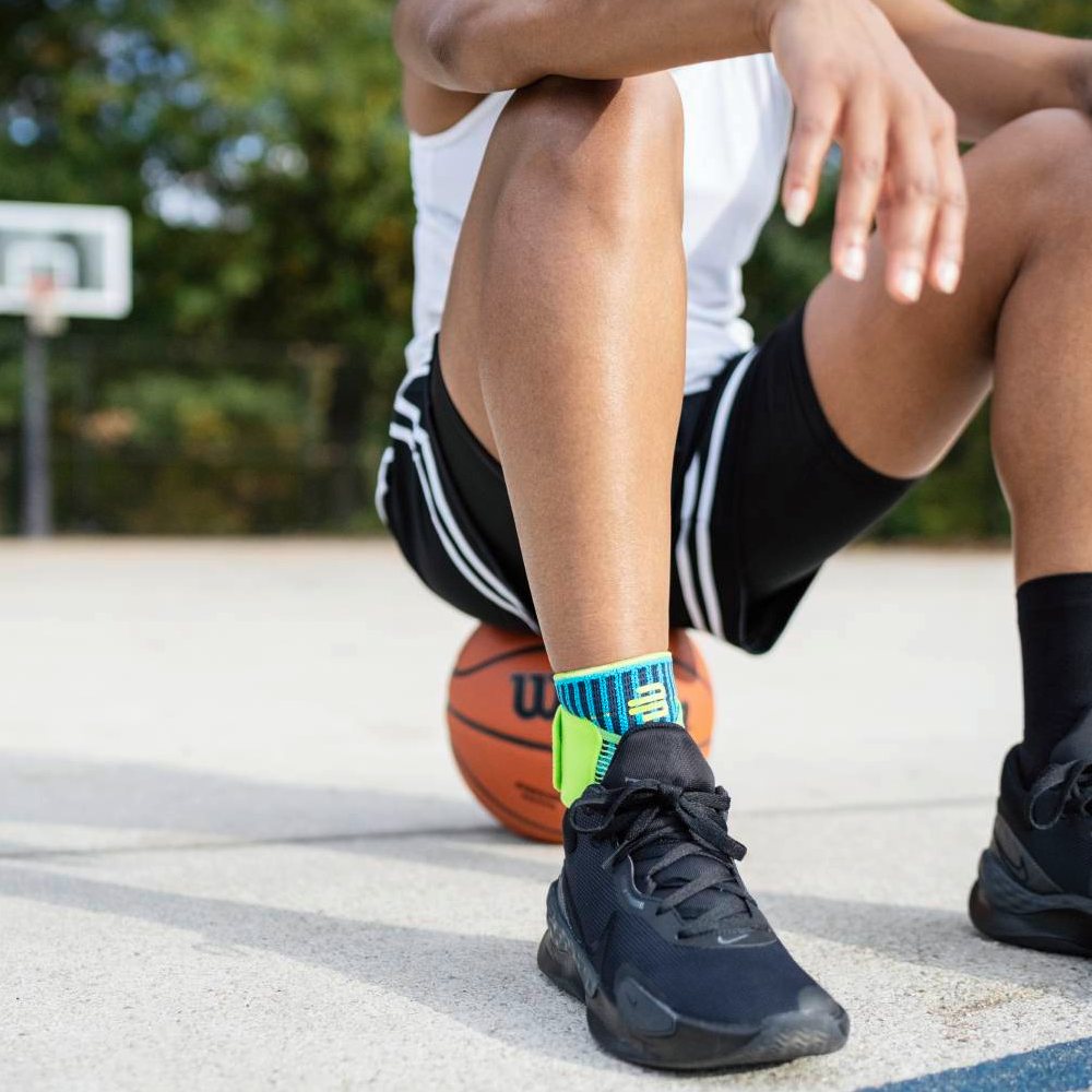 Een basketballer zit op het veld met een basketbal onder zijn been. Hij draagt de Bauerfeind Sports Ankle Support Rivera in blauw-groen om zijn enkel.