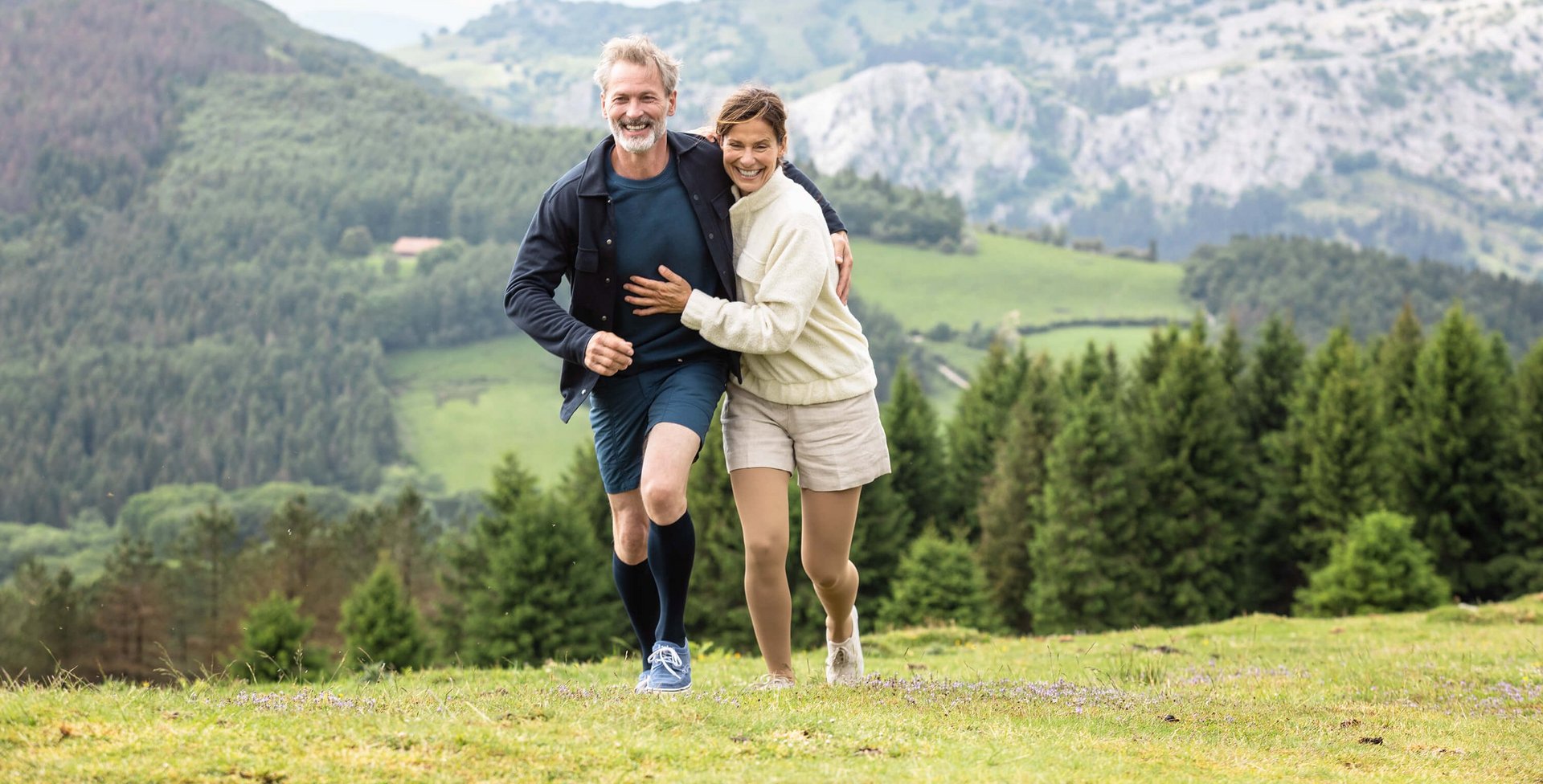 Een man en een vrouw lopen arm in arm in een berglandschap over een weide. Beiden dragen VenoTrain impuls compressiekousen van Bauerfeind. Die helpen bij spataadertjes.