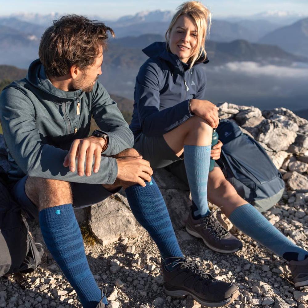 Twee wandelaars genieten van het uitzicht op een bergtop, terwijl ze Merino Outdoor compressiesokken in rood-bruin dragen. De sokken bieden optimaal comfort en ondersteuning voor lange tochten in uitdagend terrein.