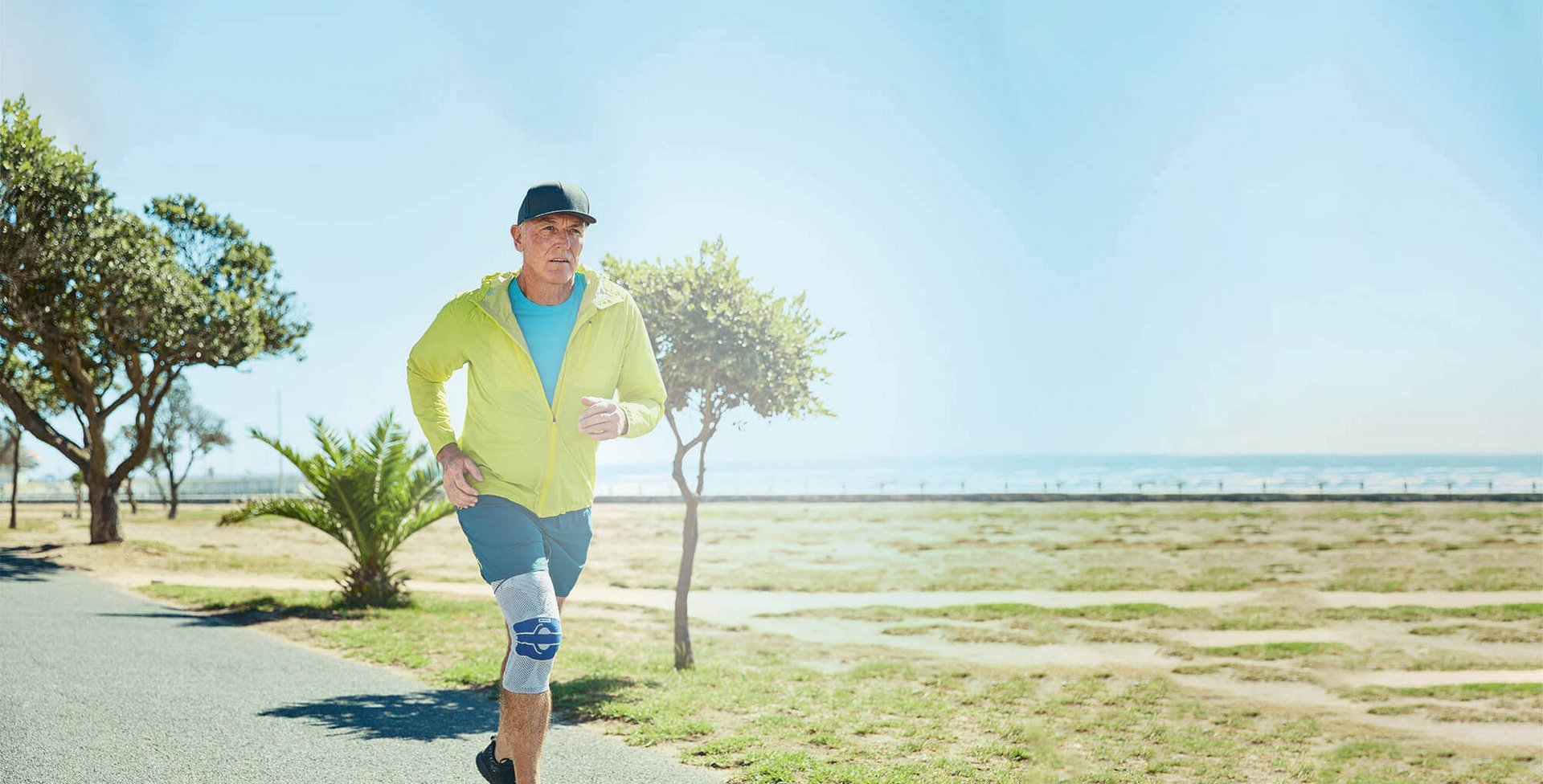Ein älterer Mann joggt auf einem sonnigen Weg entlang eines Parks in der Nähe des Meeres. Er trägt eine GenuTrain A3 Kniebandage am rechten Knie, ein gelbes Sportjackett, ein blaues T-Shirt, blaue Shorts und schwarze Laufschuhe.