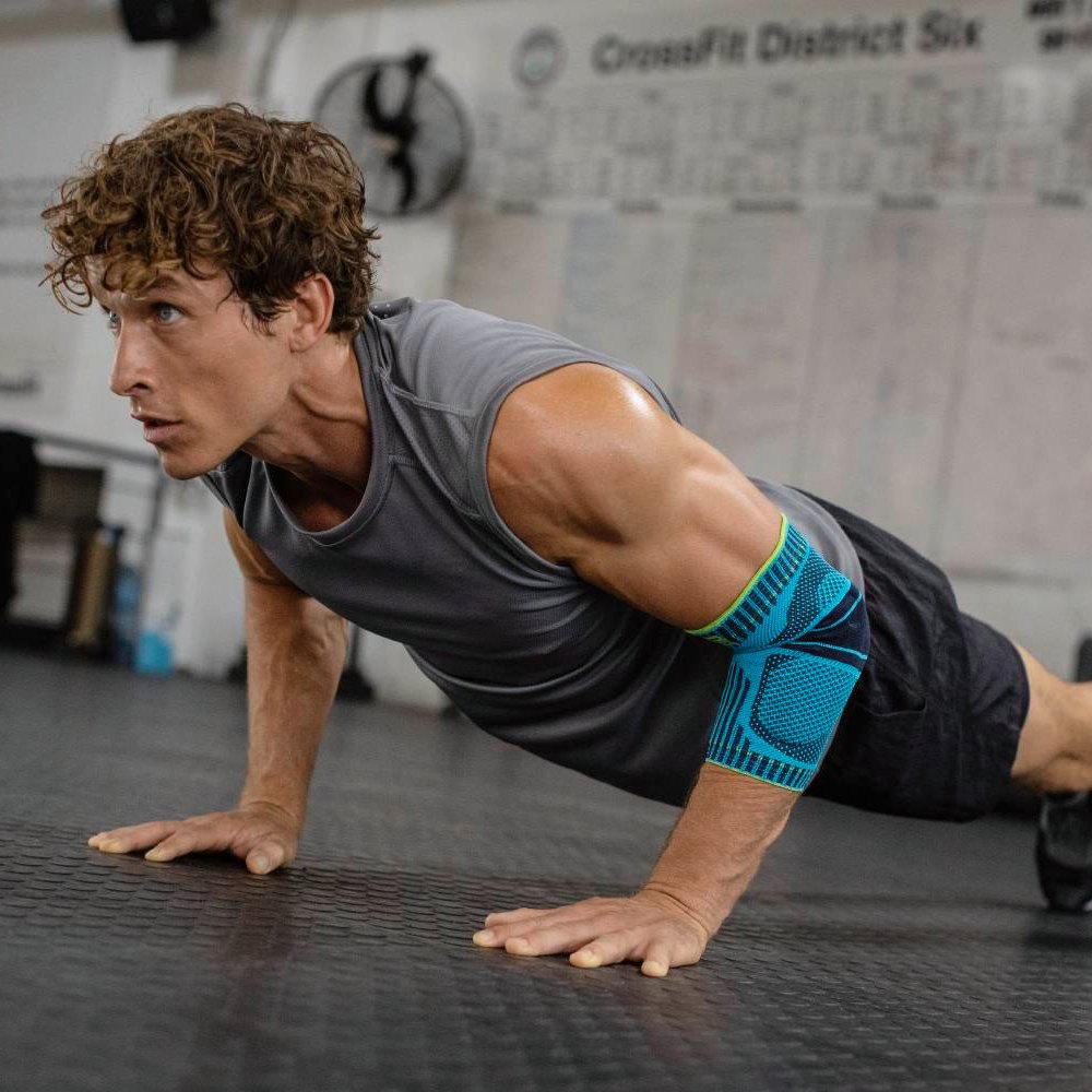 Een sportieve man met een gespierd bovenlichaam voert gefocust push-ups uit in een sportschool. Hij draagt een blauw-zwarte elleboogbandage aan zijn rechterarm voor stabiliteit en ondersteuning tijdens zijn training.