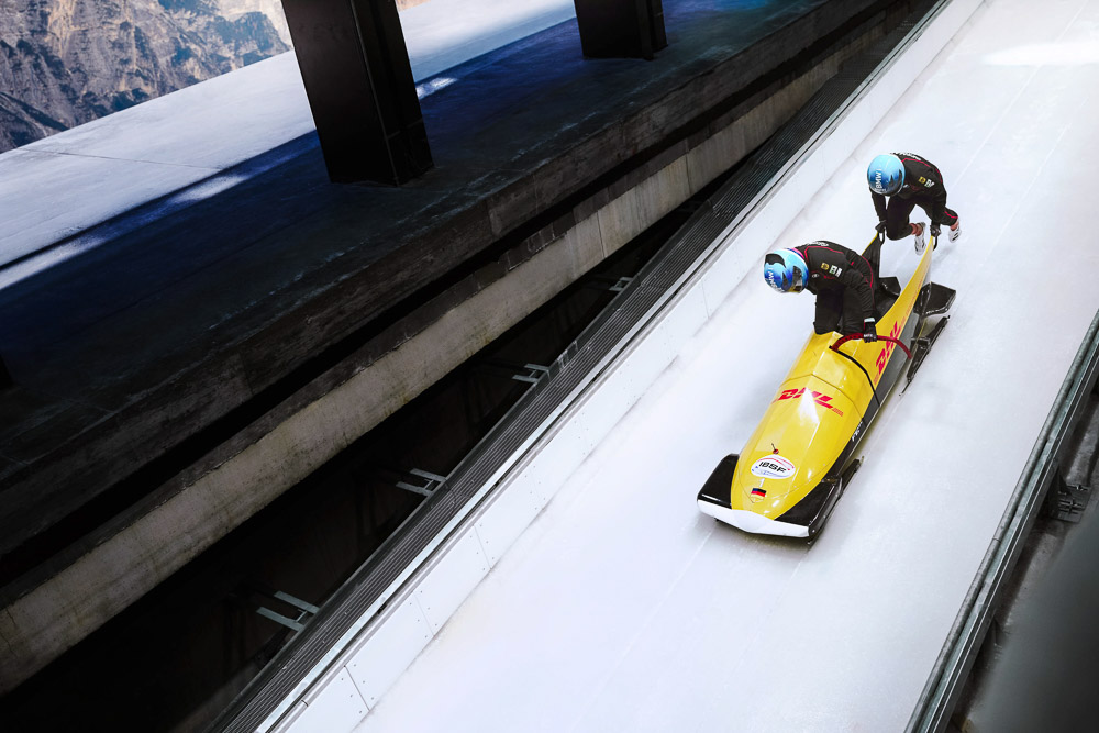 Twee bobslee-atletes duwen samen een gele tweemansbob over een ijzige baan, gefotografeerd vanuit een verhoogd perspectief. De sporters dragen helmen en strakke wedstrijdpakken, met de ijsbaan tussen betonnen wanden in bergachtig landschap.