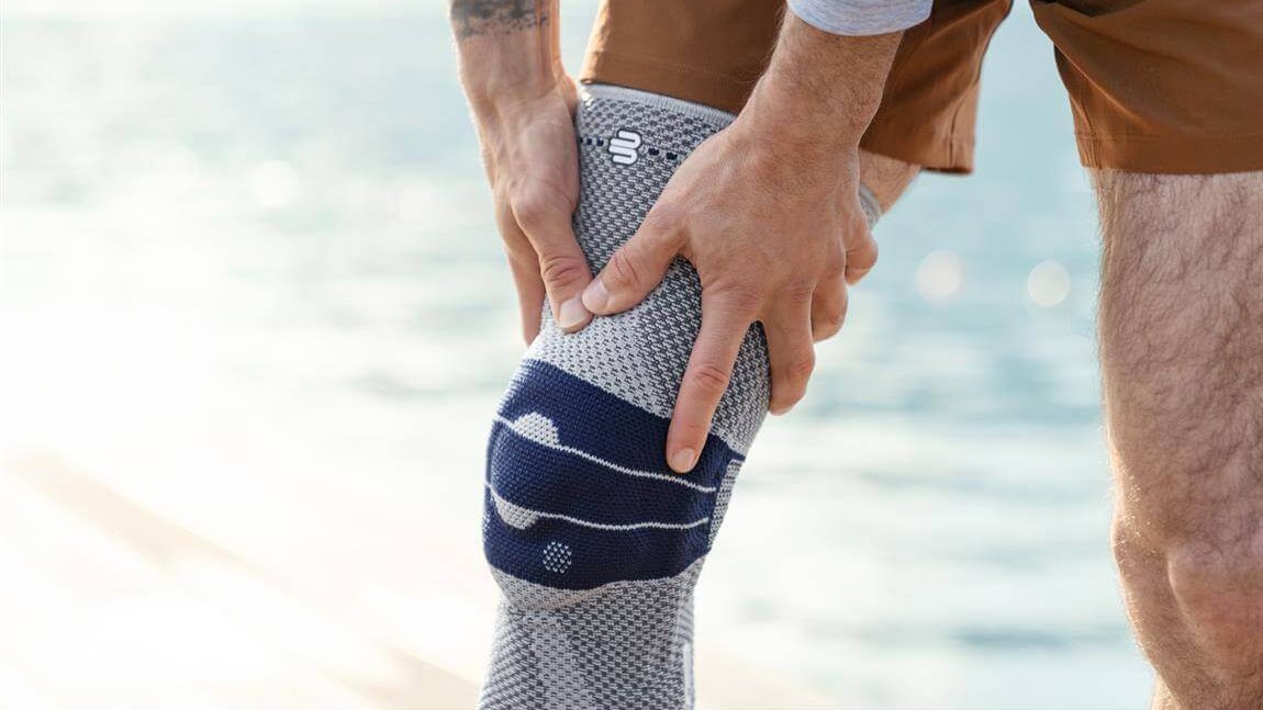 Close-up van de GenuTrain kniebandage. De bandage omsluit de rechterknie en heeft een modern, functioneel design. Op de achtergrond is wazig een waterpartij te zien.
