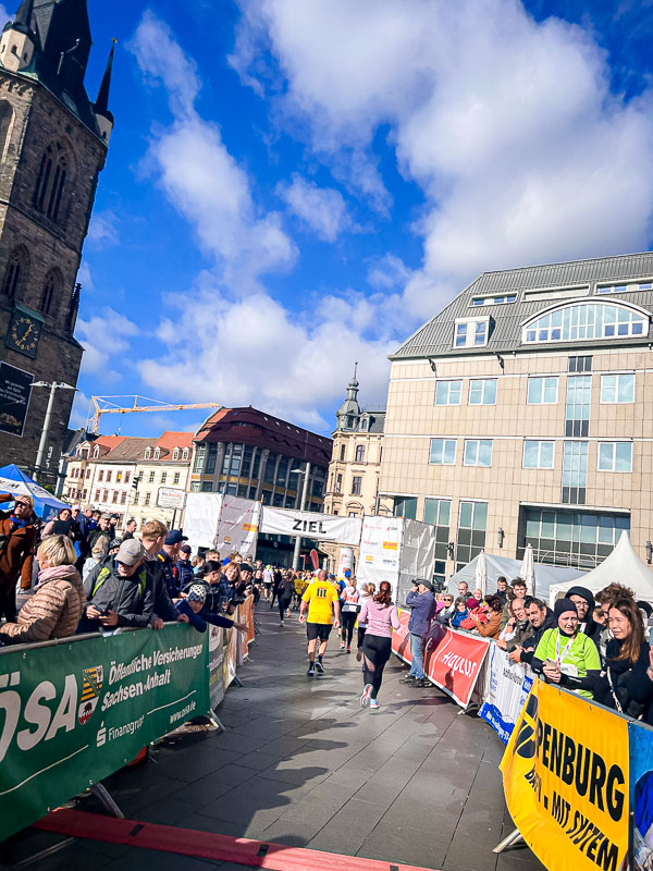 De laatste meters voor de finish bij de halve marathon in Halle. Lopers passeren de finishlijn onder een blauwe hemel, toegejuicht door het publiek.