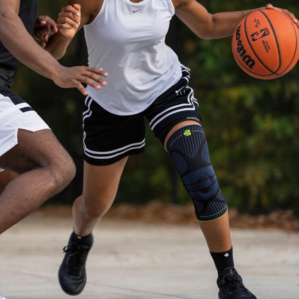 Een vrouwelijke basketbalspeelster draagt een zwarte kniebandage op haar rechterbeen terwijl ze met de bal langs een tegenstander beweegt.