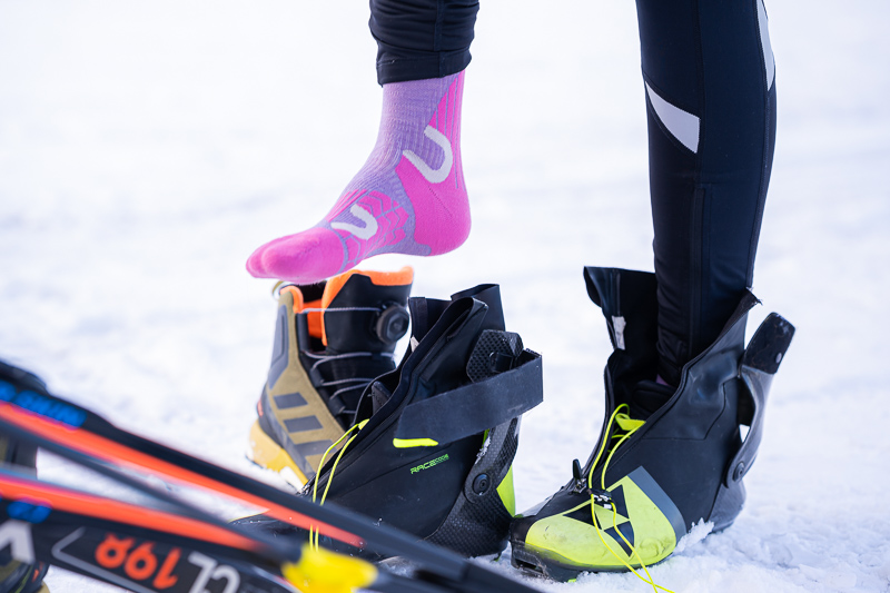 Een close-up van een langlaufuitrusting: Een atlete trekt neonroze functionele sokken aan, terwijl naast haar hoogwaardige skischoenen en ski's in de sneeuw liggen.