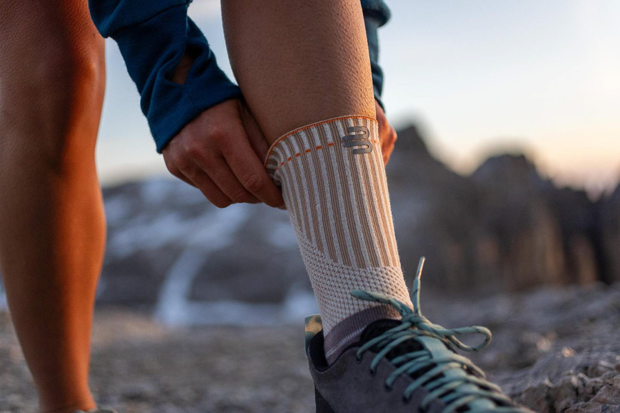Een wandelaar past zijn speciale outdoor-bandage met versterkte boord aan. Op de achtergrond zijn bergen en een zonsondergang te zien.