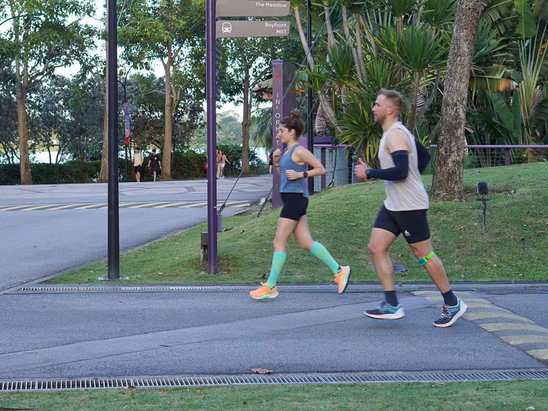 Twee mensen joggen door een stedelijk park op een geasfalteerde weg. Een man in lichte hardloopkleding en een kniebrace rent samen met een jonge vrouw die groene compressiekousen draagt.