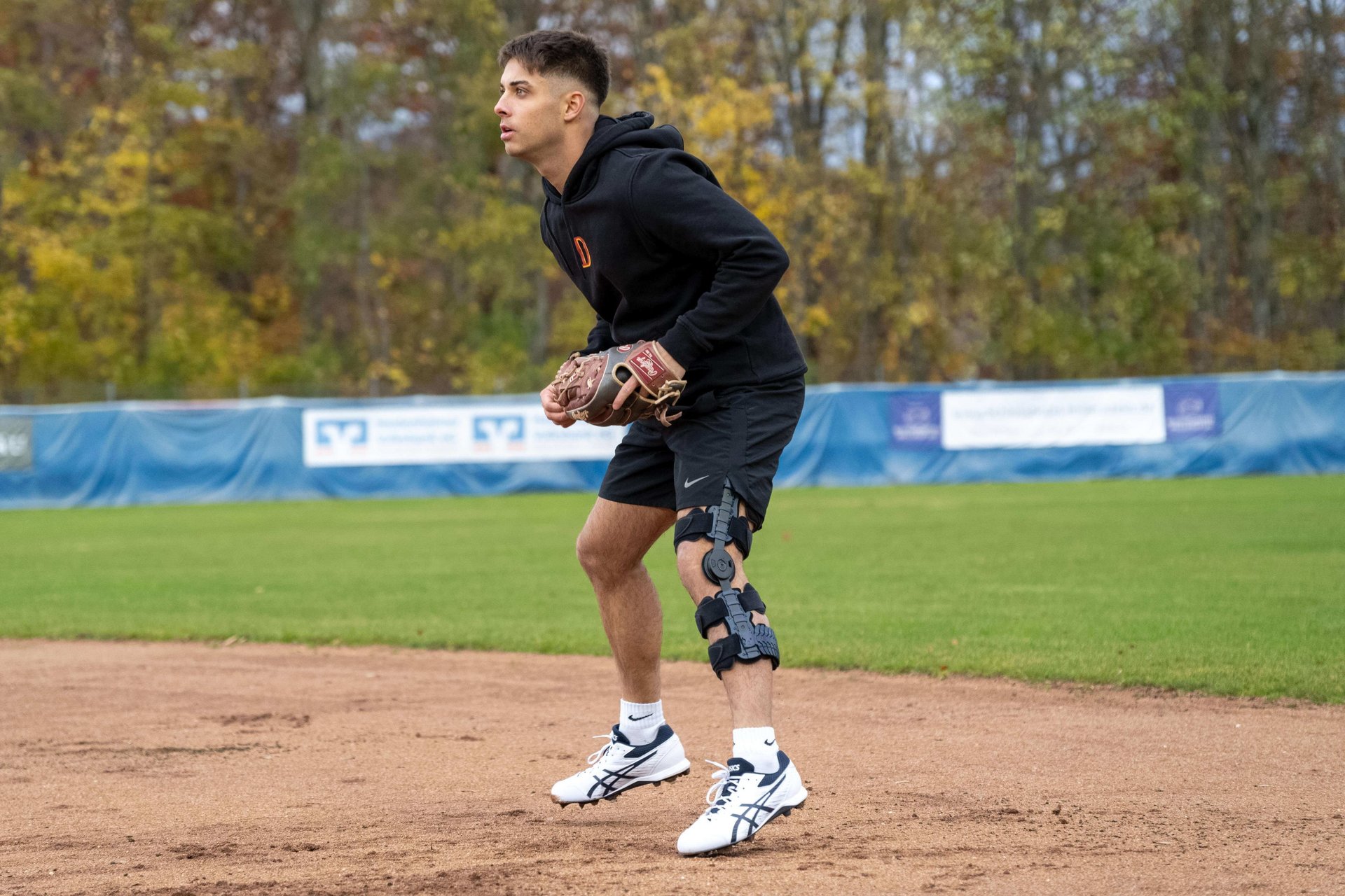 Simon Liedtke staat in de veldverdedigingspositie op een honkbalveld. Hij draagt een zwarte hoodie, een short en de SecuTec Genu Flex knieorthese om zijn rechterbeen, om zijn knie na een gescheurde achterste kruisband te stabiliseren.