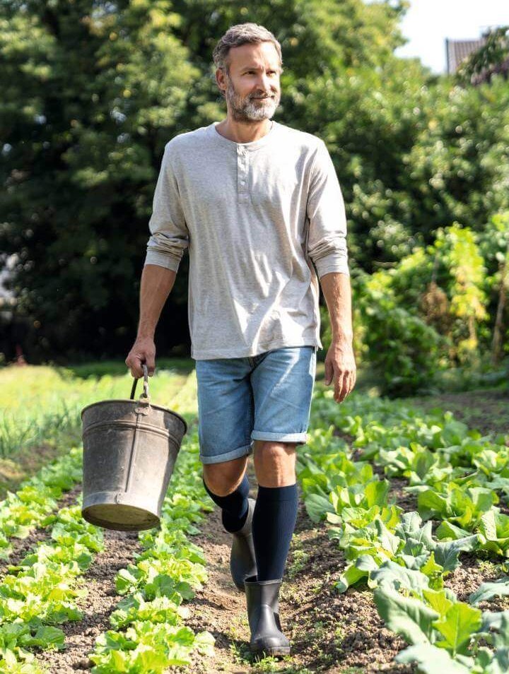 Een man in donkerblauwe VenoTrain hero compressiekousen werkt in de tuin. Hij houdt een metalen emmer vast en draagt rubberlaarzen, een grijze longsleeve en een spijkershort; op de achtergrond groene planten en bomen.