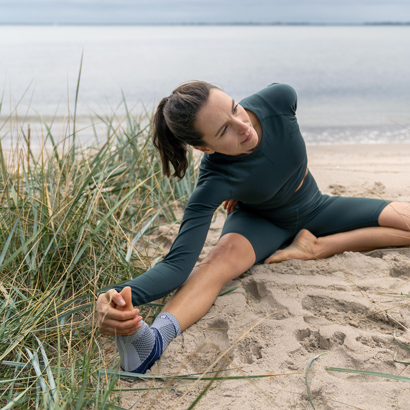 Vrouw doet yoga op zandstrand aan rustig meer met groene sportoutfit en enkelbrace.