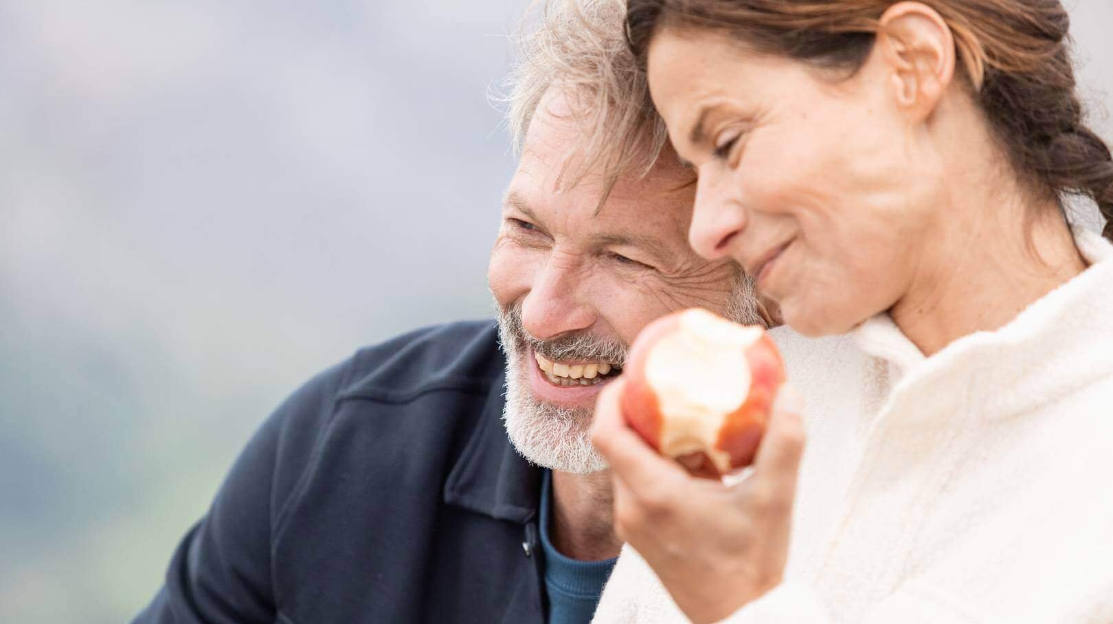 De foto toont een man en een vrouw. De vrouw eet net een appel terwijl de man lacht.