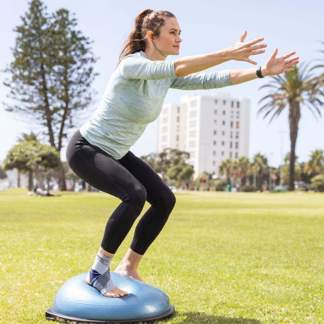 De afbeelding toont een vrouw bij een fysiotherapeutische oefening op een balanceboard in het park. Ter ondersteuning van haar enkel draagt ze een MalleoTrain bandage van Bauerfeind.
