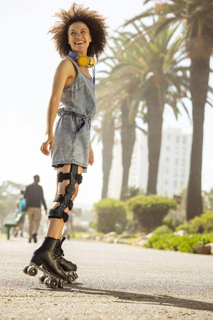 De afbeelding toont een lachende, jonge vrouw met rolschaatsen op een strandpromenade. Aan de knie draagt ze een SecuTec Genu. De orthese biedt onder andere ondersteuning na een kruisbandscheur.