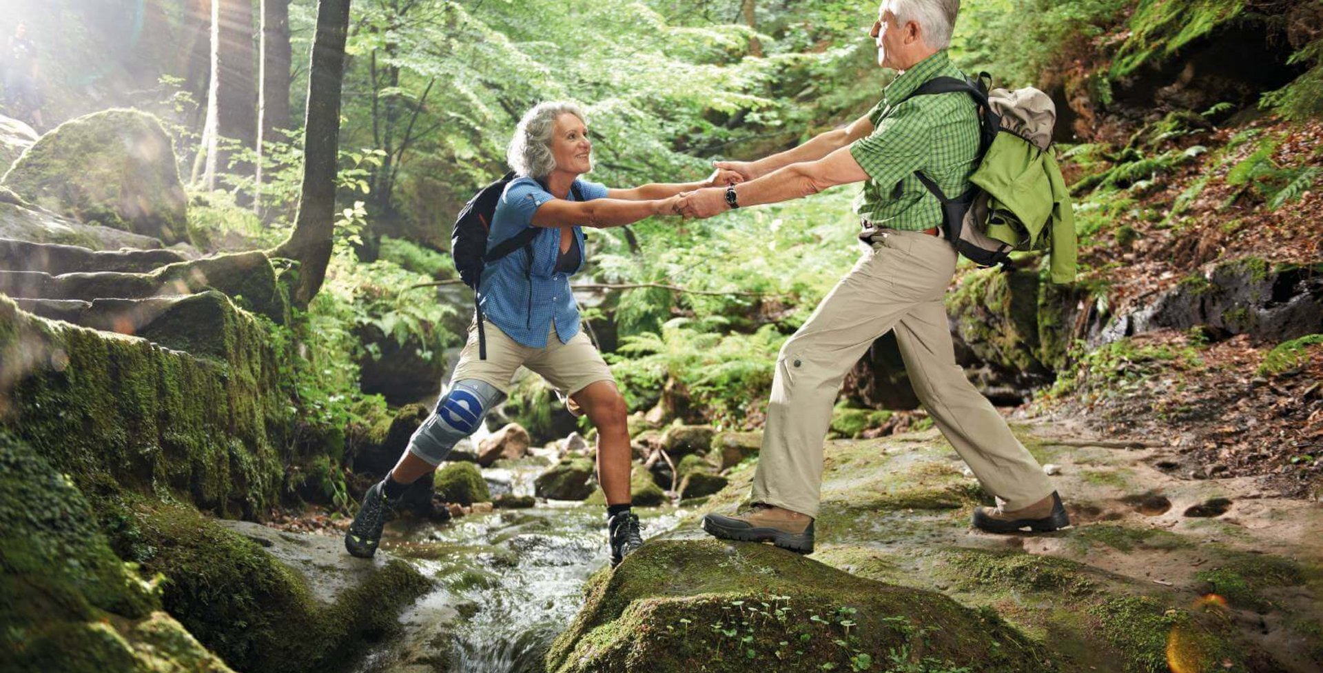 De afbeelding toont een echtpaar tijdens een trektocht in het bos. De man helpt zijn vrouw over een rivier. Aan de knie draagt de wandelaarster een SoftTec Genu van Bauerfeind ter ondersteuning.