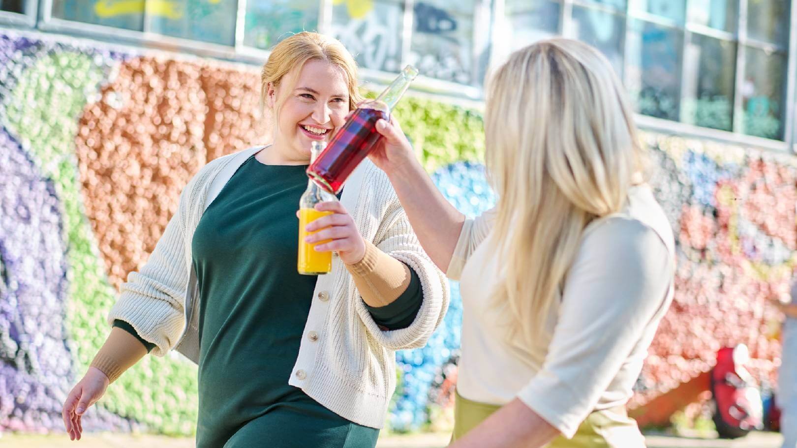 Twee vrouwen klinken met kleurrijke drinkflessen, terwijl ze lachend door een stedelijke omgeving wandelen. Op de achtergrond is een kleurrijke muur met grafische patronen te zien, die een levendige sfeer schept.