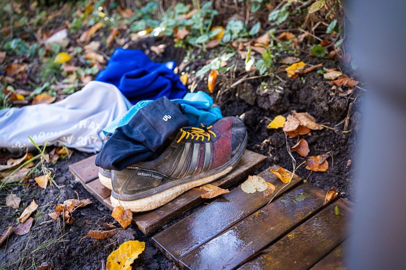 Een paar hardloopschoenen met gele veters op een houten plank. Ernaast ligt een stapel kleding met een blauwe handdoek, een grijs jack en zwarte handschoenen. Herfstbladeren liggen verspreid op de grond.