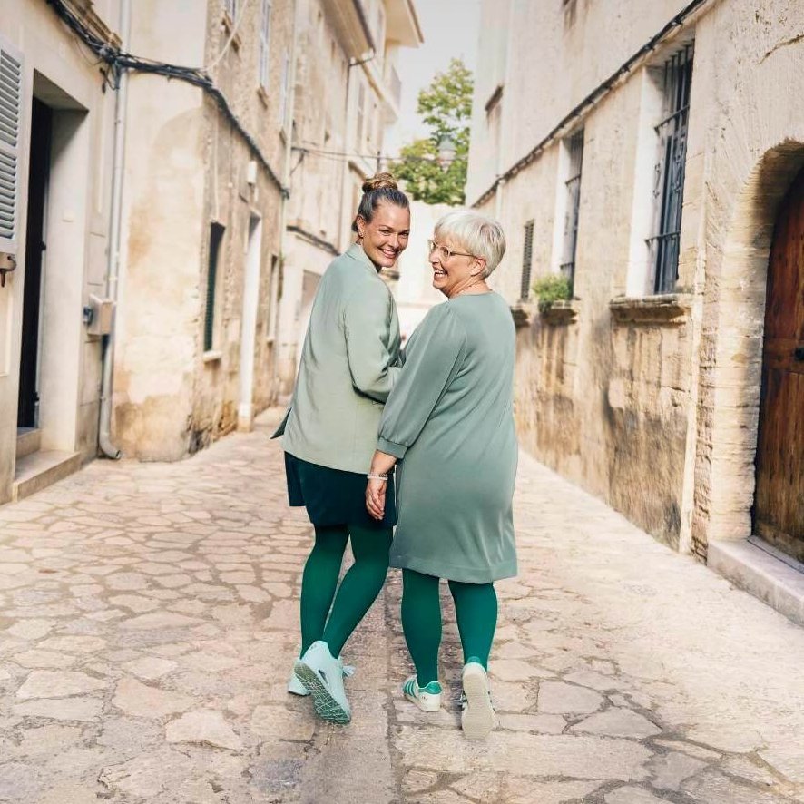 Twee vrouwen, een jongere en een oudere, wandelen lachend hand in hand door een charmant, smal straatje in de oude binnenstad. Beiden dragen qua kleur op elkaar afgestemde outfits in groen met bijpassende compressie en sneakers.