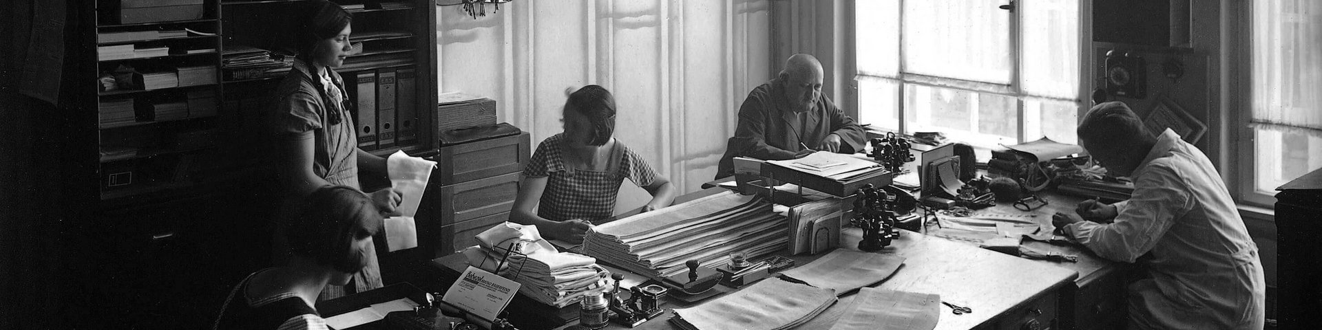 Zwart-witfoto van een oud kantoor met vijf personen die aan een grote tafel werken. Er hangt een drukke sfeer met typemachines, stapels papier en hanglampen. Op de achtergrond zie je grote ramen en een archiefkast met talloze dozen.