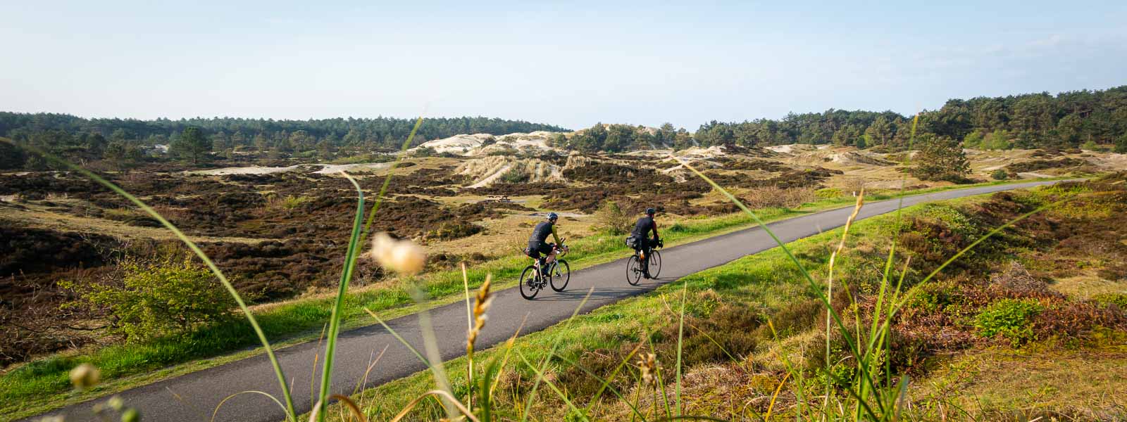 Twee fietsers die meedoen aan de Race om Nederland rijden over een weg door de duinen.