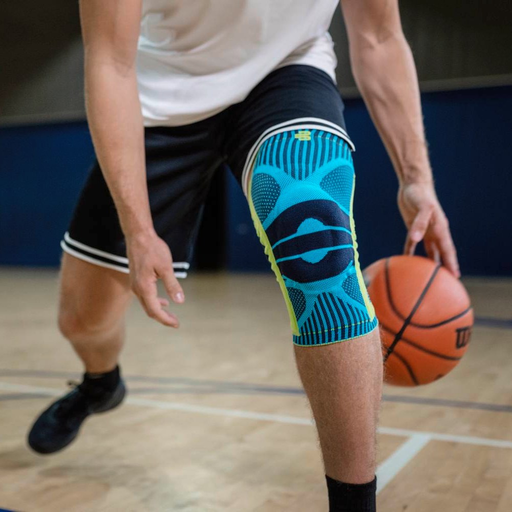 Een basketbalspeler met een turquoise kniebandage op zijn rechterbeen dribbelt de bal in een sporthal; de nadruk ligt op het product en zijn houding.