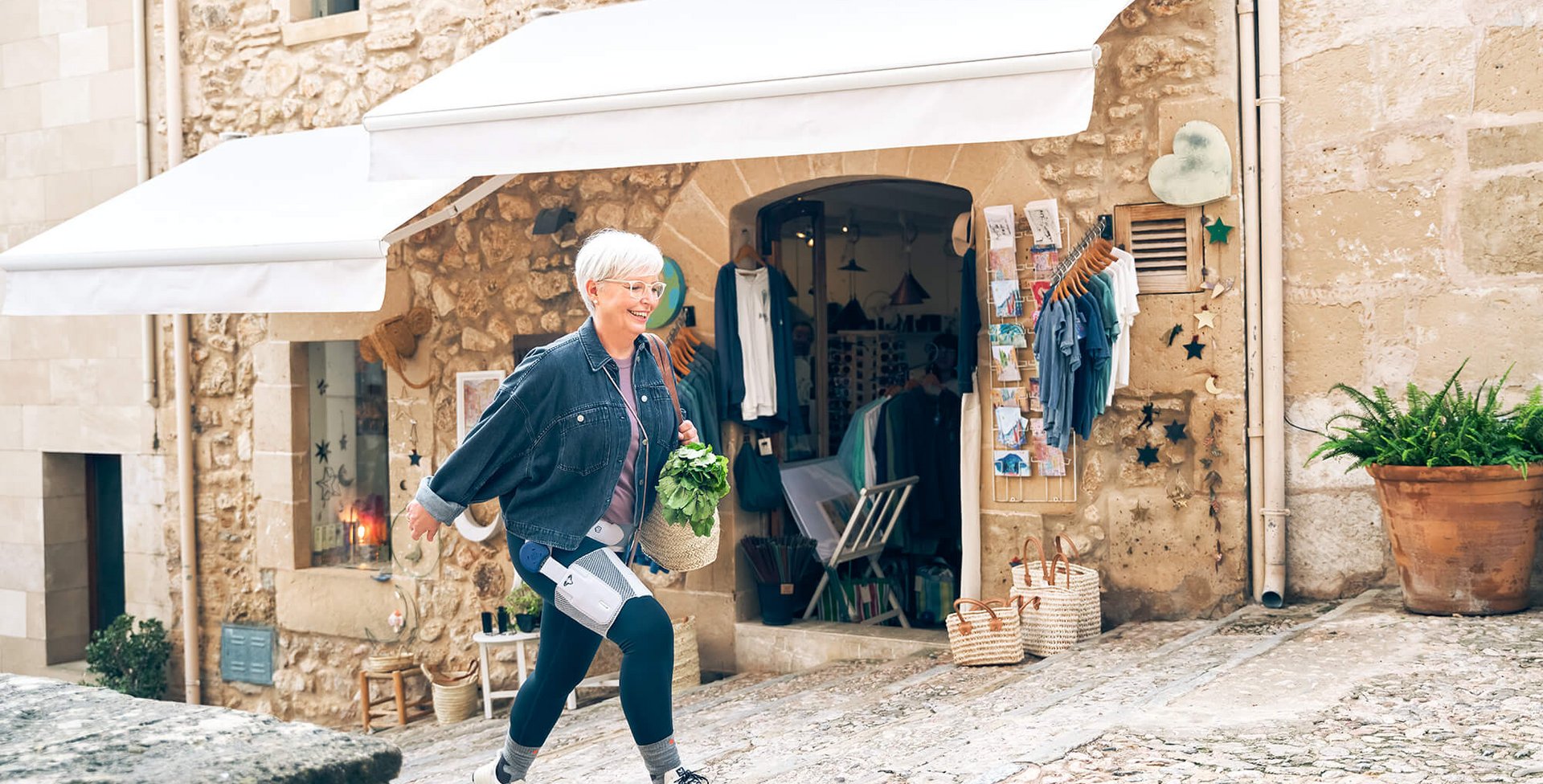 Een oudere vrouw met kort wit haar en bril loopt een geplaveide steeg omhoog. Ze draagt een spijkerjasje en een donkere legging met medische hulpmiddelen en houdt een mand met verse groenten vast. Op de achtergrond een winkel met witte zonneschermen.