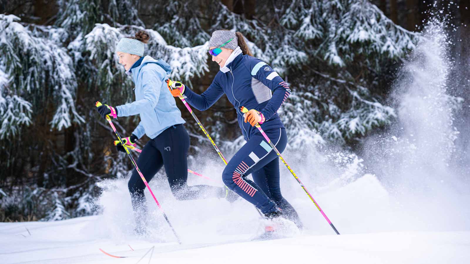 Twee vrouwen tijdens het langlaufen door met sneeuw bedekte bossen. Ze dragen kleurrijke wintersportkleding en glijden dynamisch door de sneeuw. De scène wordt gekenmerkt door dicht met sneeuw bedekte sparren, die het winterwonderland benadrukken.