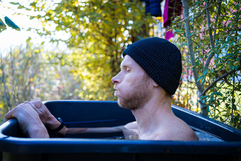 Een man met een donkere gebreide muts zit tot aan zijn schouders in een ijsbad in de buitenlucht. Hij is omringd door herfstige vegetatie en zijn gezichtsuitdrukking toont diepe ademhaling om zich aan de kou aan te passen.