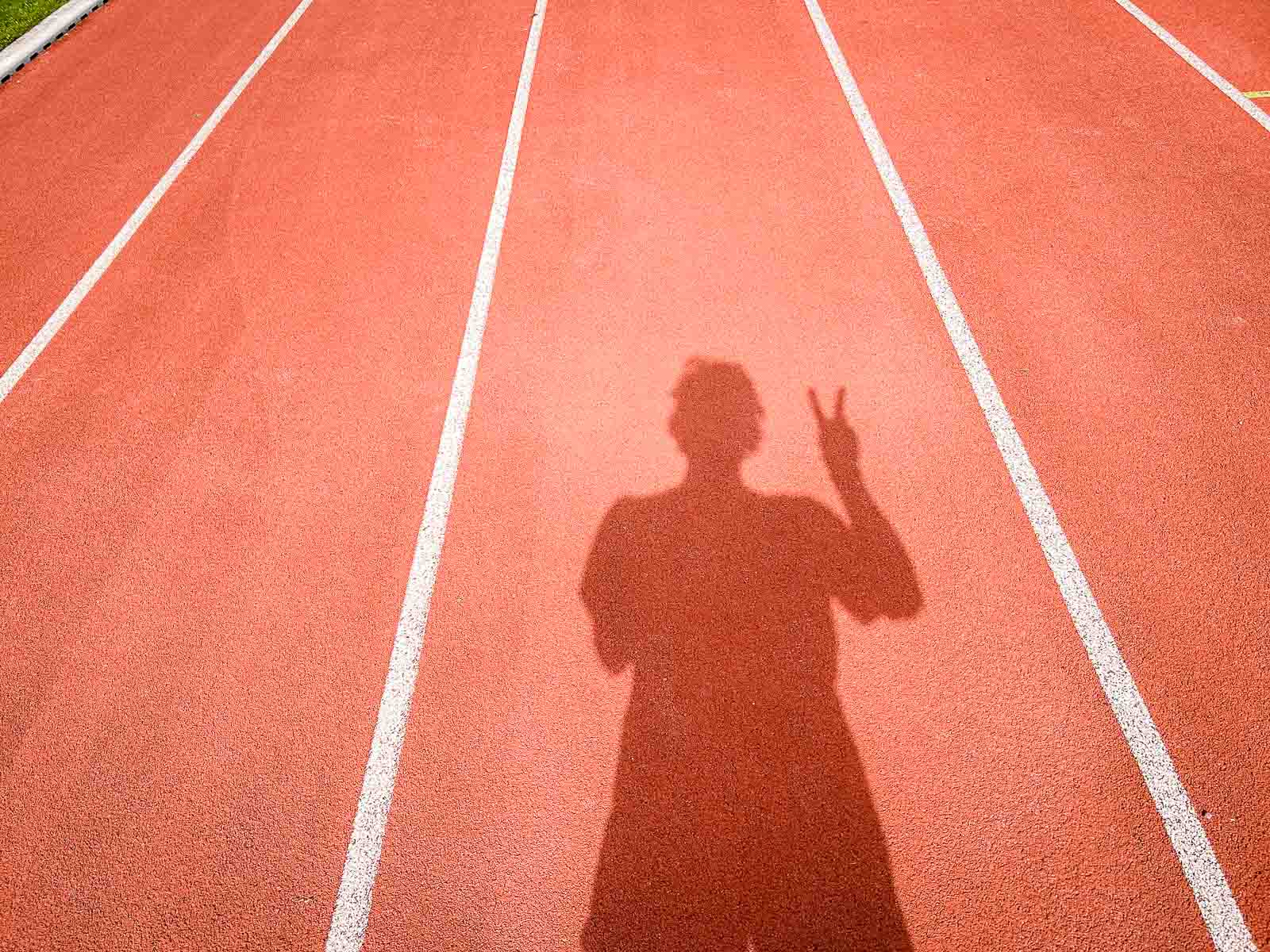 De schaduw van een loper op een rode renbaan die het victory-teken met zijn hand toont. De scène symboliseert succes en het behalen van doelen tijdens hardlooptraining.