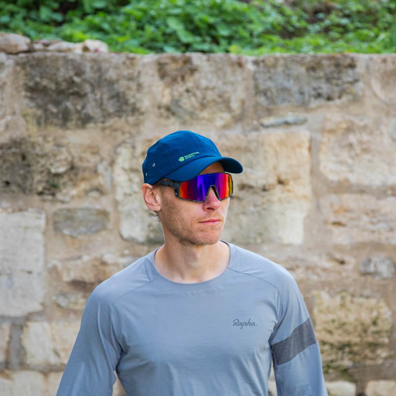 Sporter draagt een blauwe Bauerfeind Sports pet en spiegelende sportbril voor een stenen muur – klaar voor actieve momenten buiten.