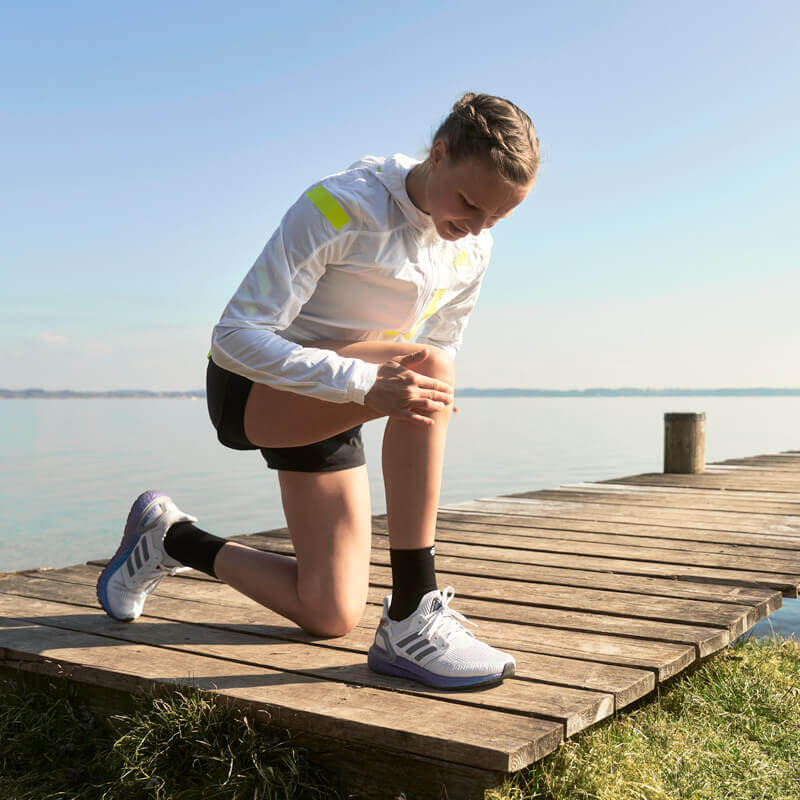 De afbeelding toont hardloopster Anna Hahner, die aan de oever van een meer op een houten steiger haar knie onderzoekt. Ontdek wat je tegen kniepijn tijdens het lopen kunt doen.