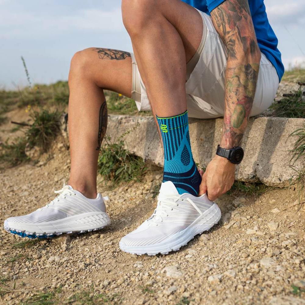 Een man zit op een betonnen rand in een natuurlijke omgeving met een grindpad en gras. Hij draagt witte hardloopschoenen en blauwe sportsokken met een opvallend patroon. Zijn handen trekken de schoen over de sok terwijl zijn getatoeëerde arm zichtbaar is.