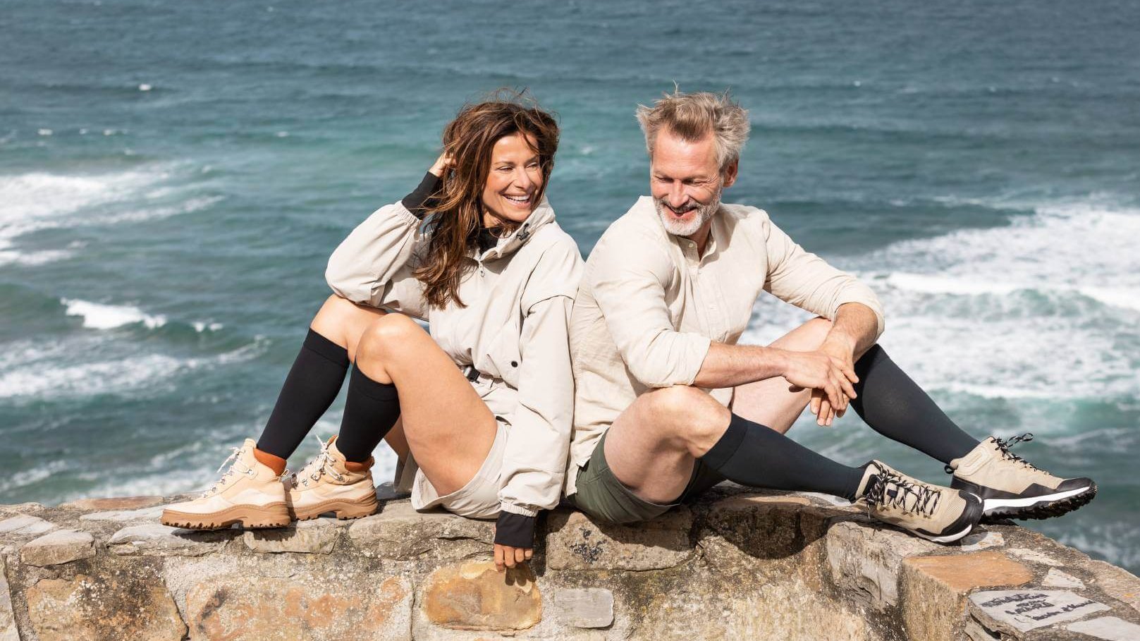Een man en een vrouw zitten ontspannen op een stenen muur met uitzicht op zee op de achtergrond. Beiden dragen outdoorkleding en zwarte compressiekousen van de lijn VenoTrain, gecombineerd met wandelschoenen.