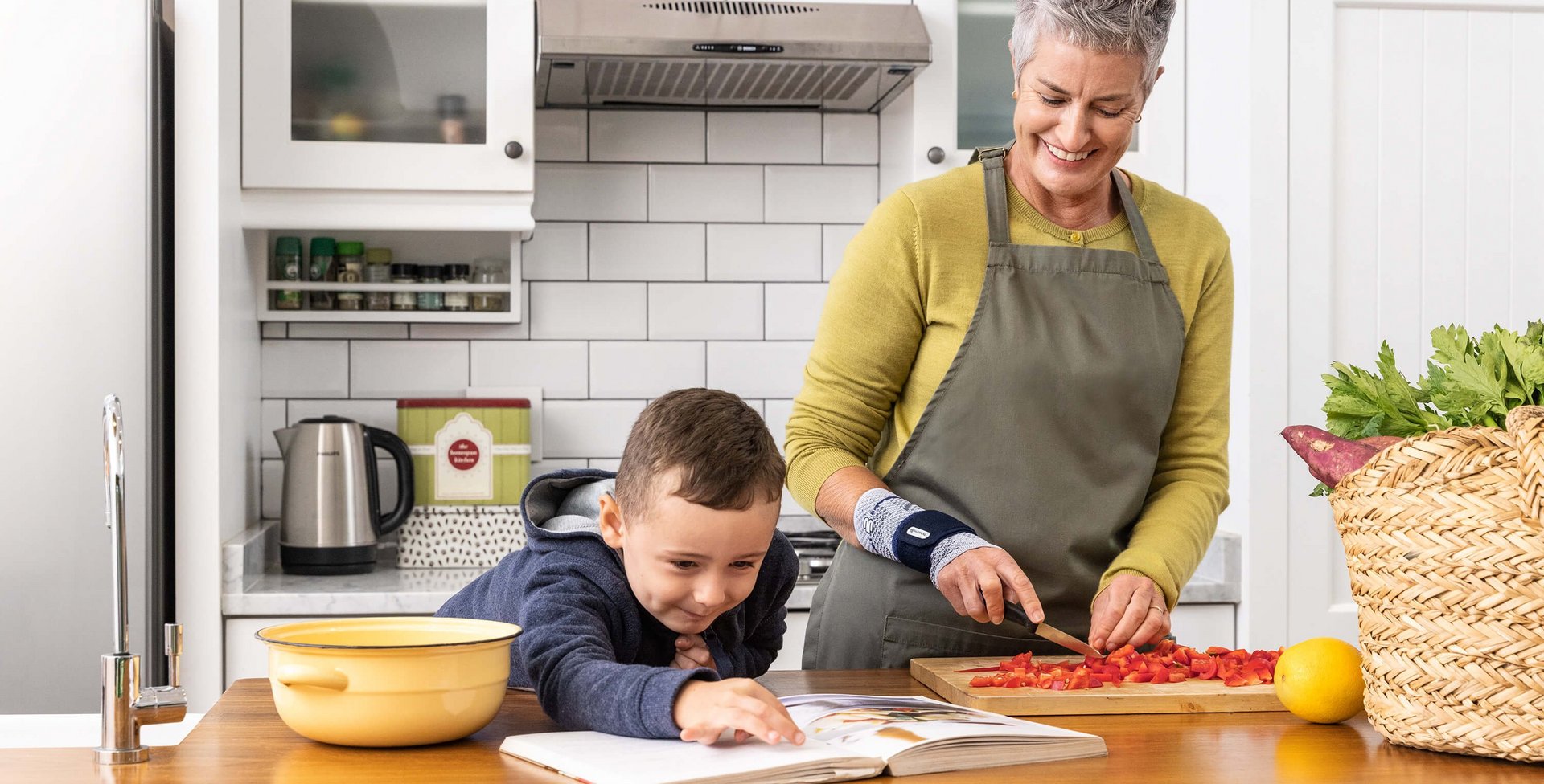 De foto toont een oudere vrouw in een keuken die samen met haar kleinzoon kookt. De dame draagt een ManuTrain bandage voor de conservatieve behandeling van haar polsartrose.