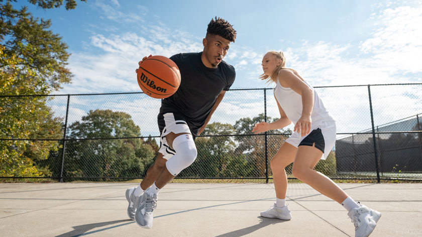 Een basketballer sprint over een buitenveld met de bal, op de hielen gezeten door een tegenstander. Hij draagt een witte kniebrace voor extra stabiliteit. De omgeving is herfstachtig met bomen en een hekwerk rondom.