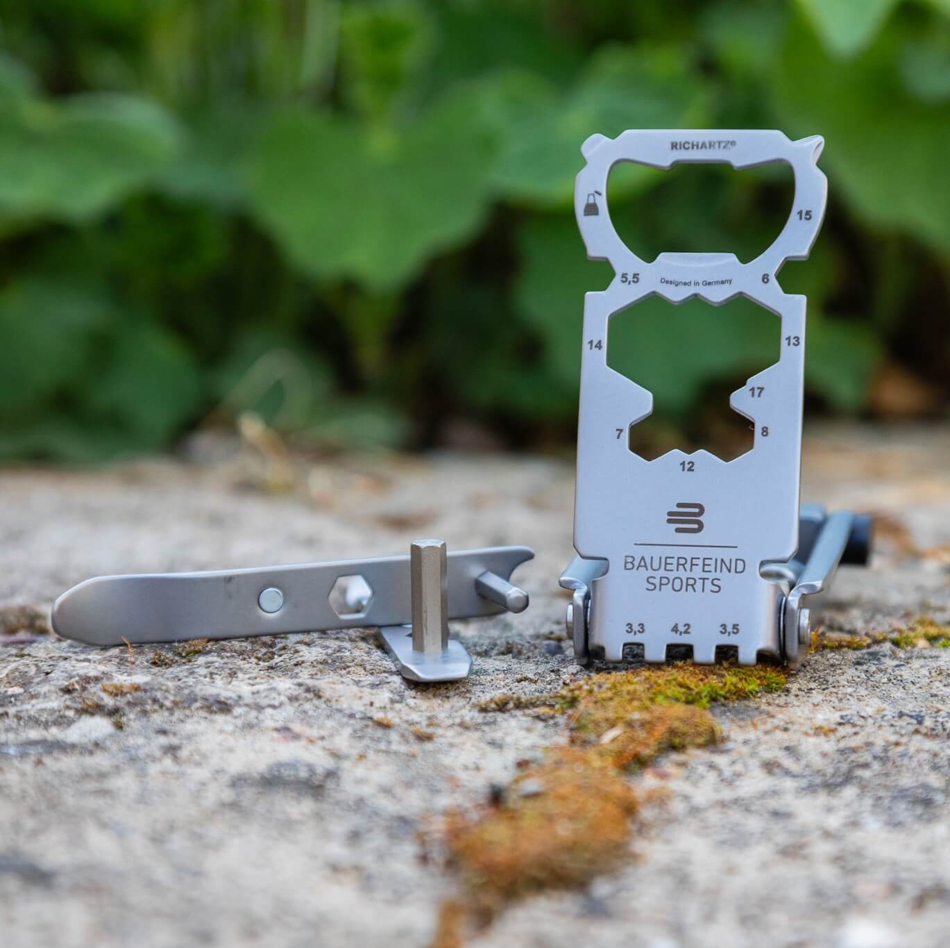 Bauerfeind Sports multitool met verschillende gereedschapsfuncties en een losse bit op een stenen ondergrond met mos, met groene planten op de achtergrond – het praktische hulpmiddel voor snelle reparaties en zorgeloze fietsmomenten onderweg.