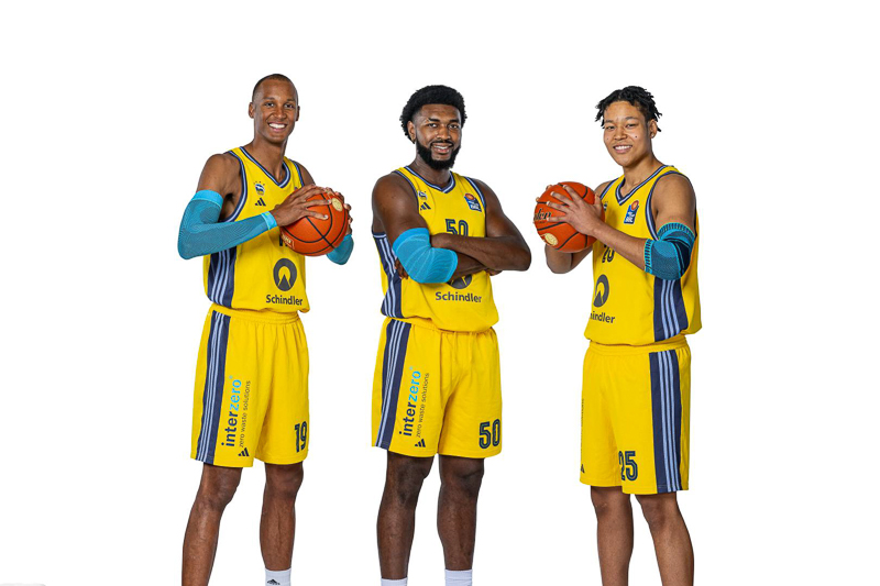 Drie basketballers van Alba Berlin in hun gele teamtenues poseren met basketbal voor een neutrale witte achtergrond. Elke speler glimlacht en houdt een basketbal vast met een ontspannen, professionele uitstraling.