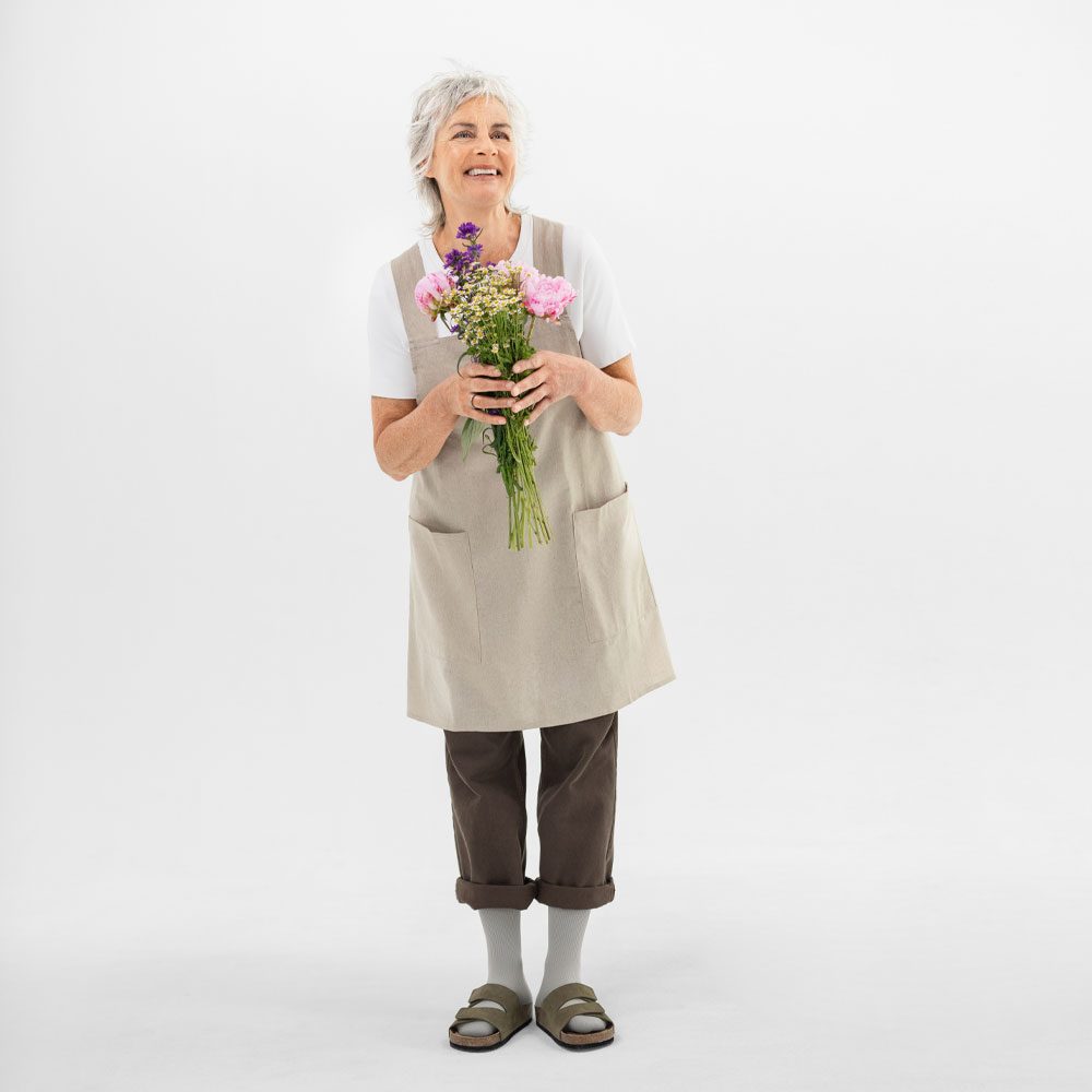 Oudere vrouw in schort houdt een boeket roze, paarse en witte bloemen vast tegen een lichte achtergrond.