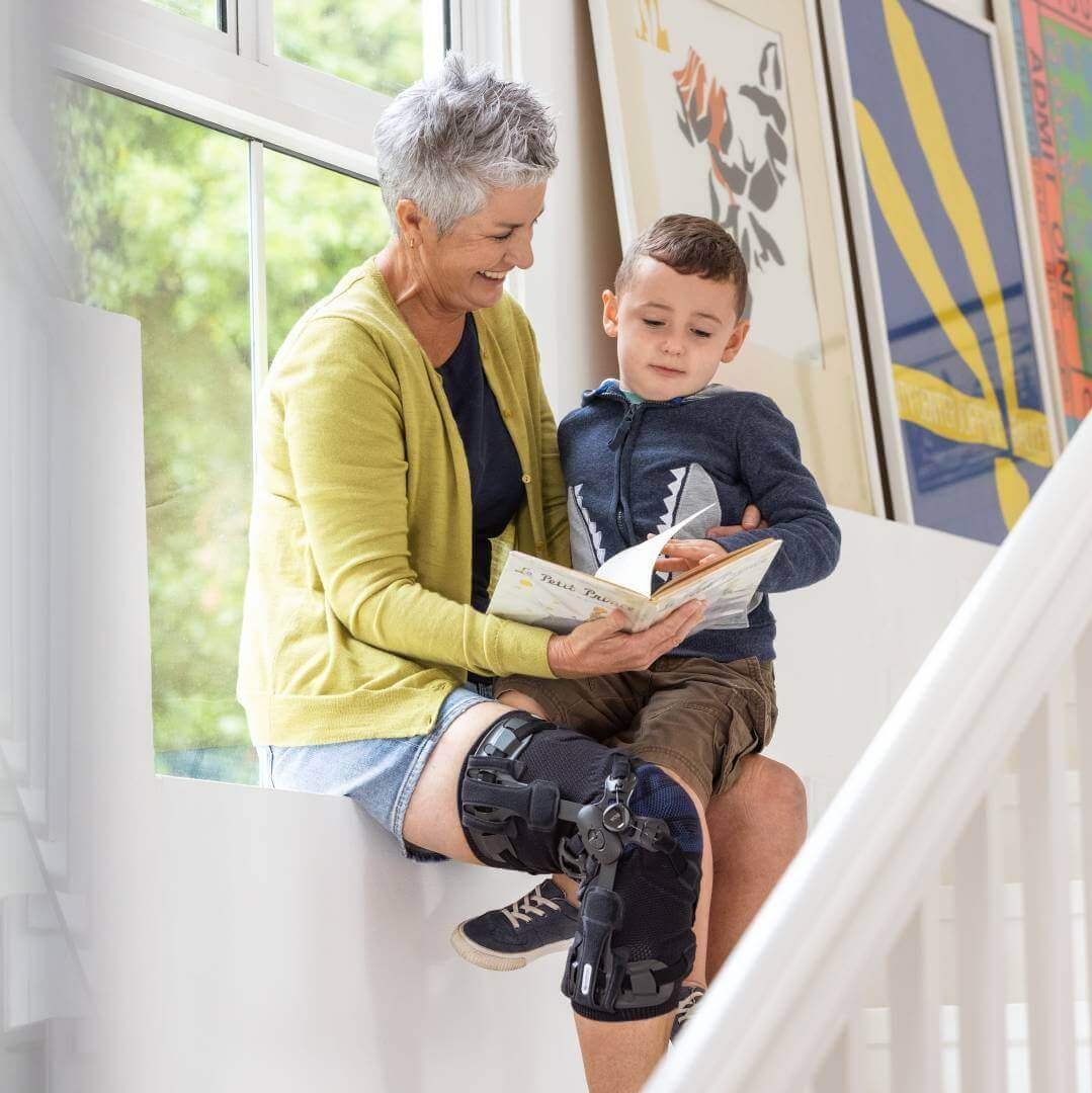 De foto toont een oudere vrouw die samen met haar kleinkind in een boek leest. De dame draagt een GenuTrain OA-orthese voor de conservatieve behandeling van artrose.