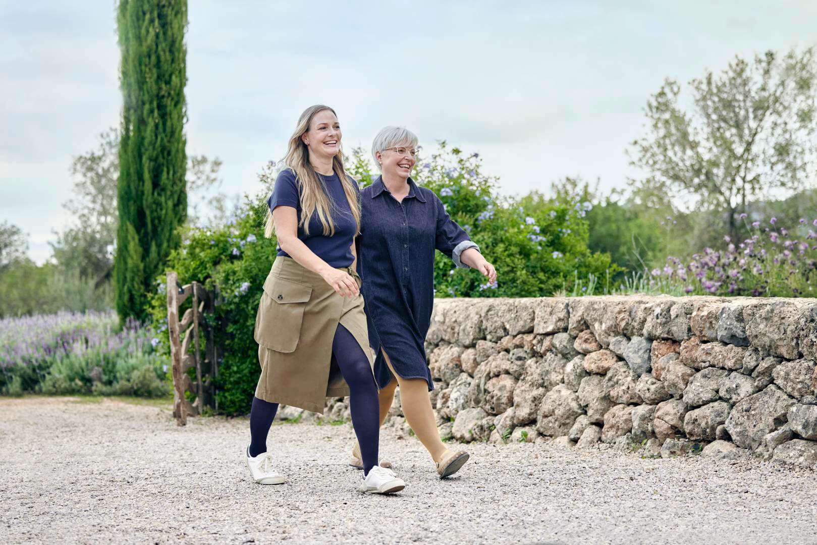 Twee vrouwen wandelen lachend over een grindpad door een idyllisch landschap met lavendel en groene struiken. Ze dragen comfortabele, stijlvolle outfits met VenoTrain compressiekousen in donkerblauw en beige, die harmonieus in hun look zijn geïntegreerd.