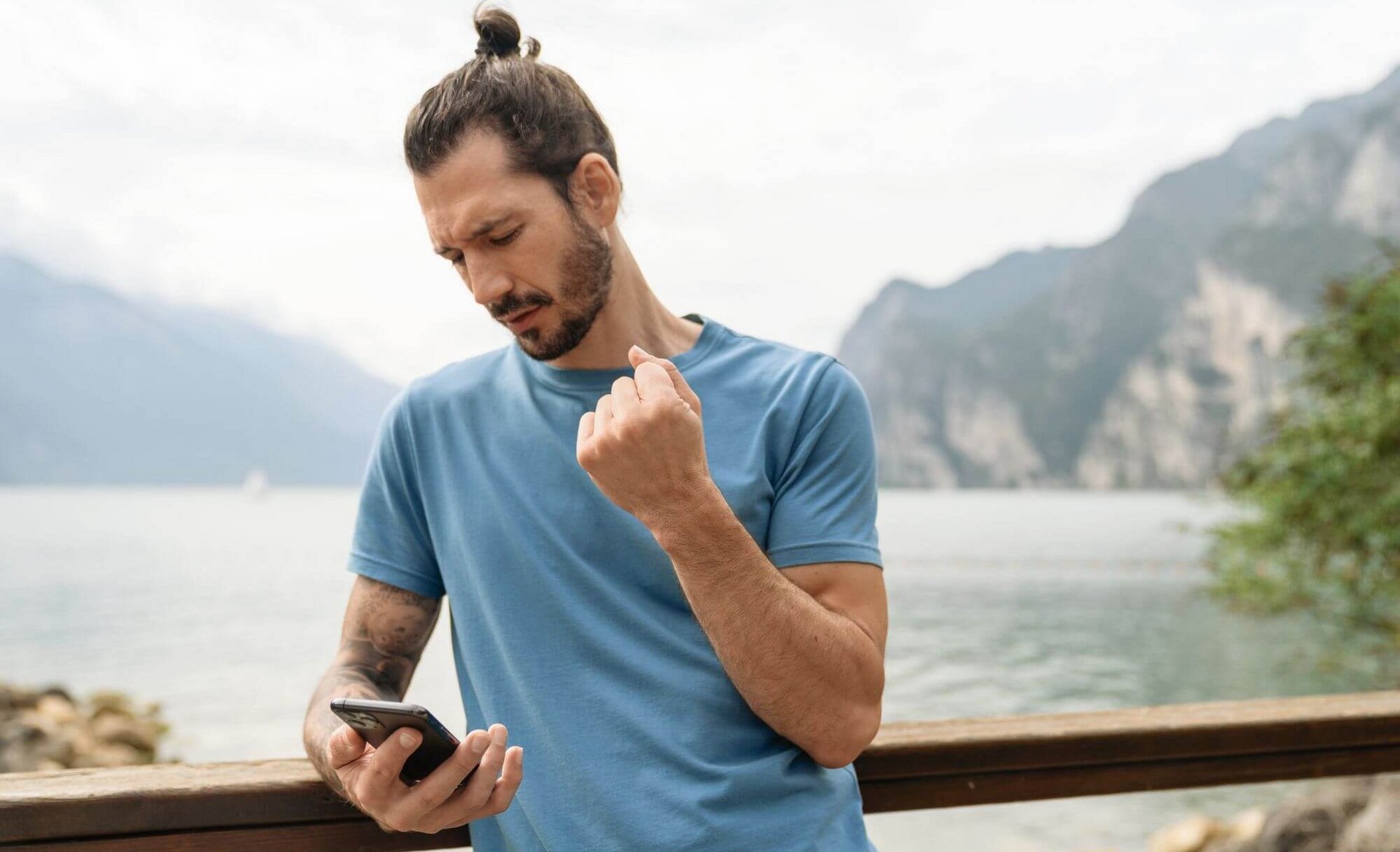 De afbeelding toont een man met smartphone die aan de oever van een bergmeer staat. Hij houdt zijn rechterarm met pijnlijke pols omhoog.