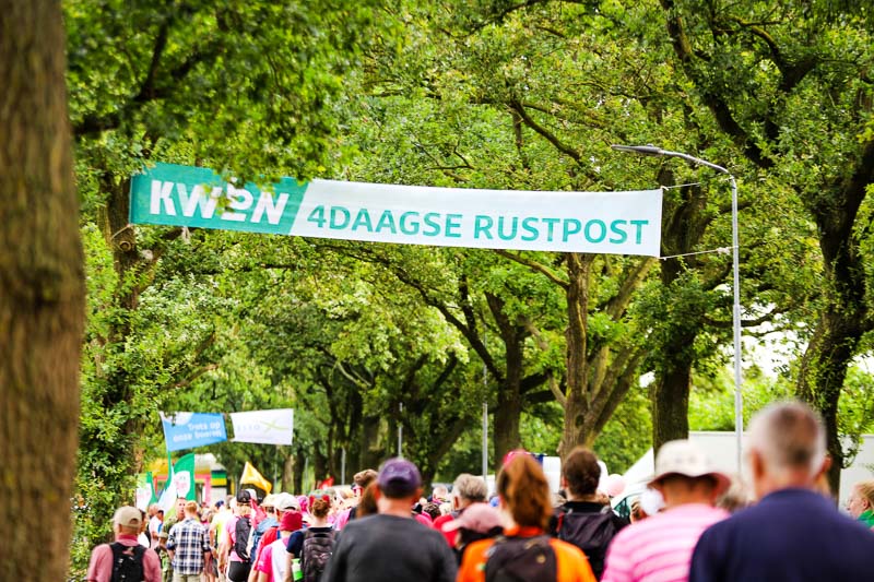 Talloze deelnemers passeren een rustpunt tijdens de 4Daagse. Boven hen hangt een groot spandoek met de tekst "KWBN 4DAAGSE RUSTPOST".