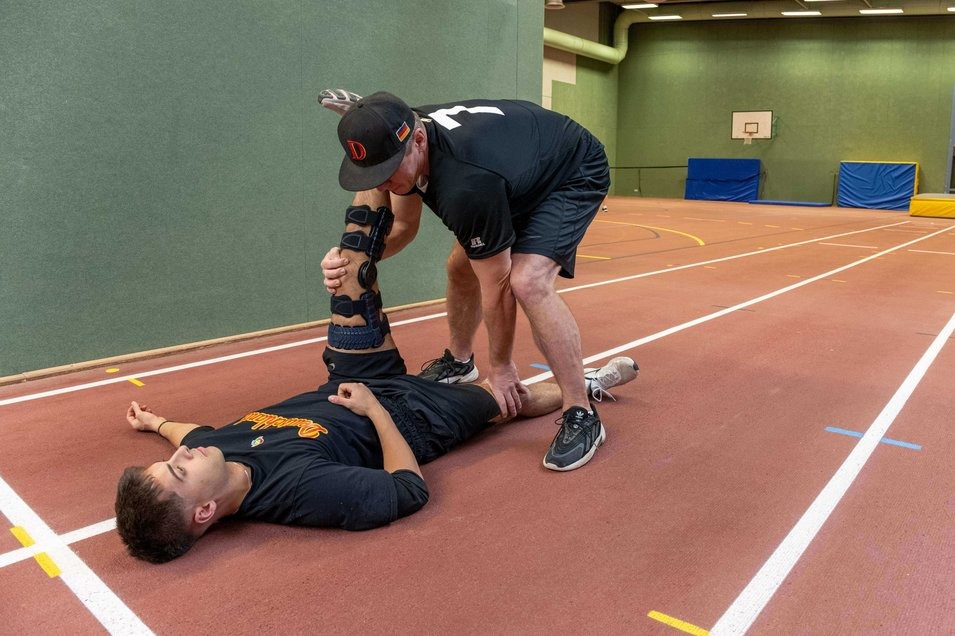 Honkballer Simon Liedtke ligt tijdens een revalidatieoefening in een sporthal op de grond. Zijn been wordt door een trainer gerekt, terwijl hij een SecuTec Genu Flex knieorthese draagt om het gewricht na een gescheurde achterste kruisband te stabiliseren.