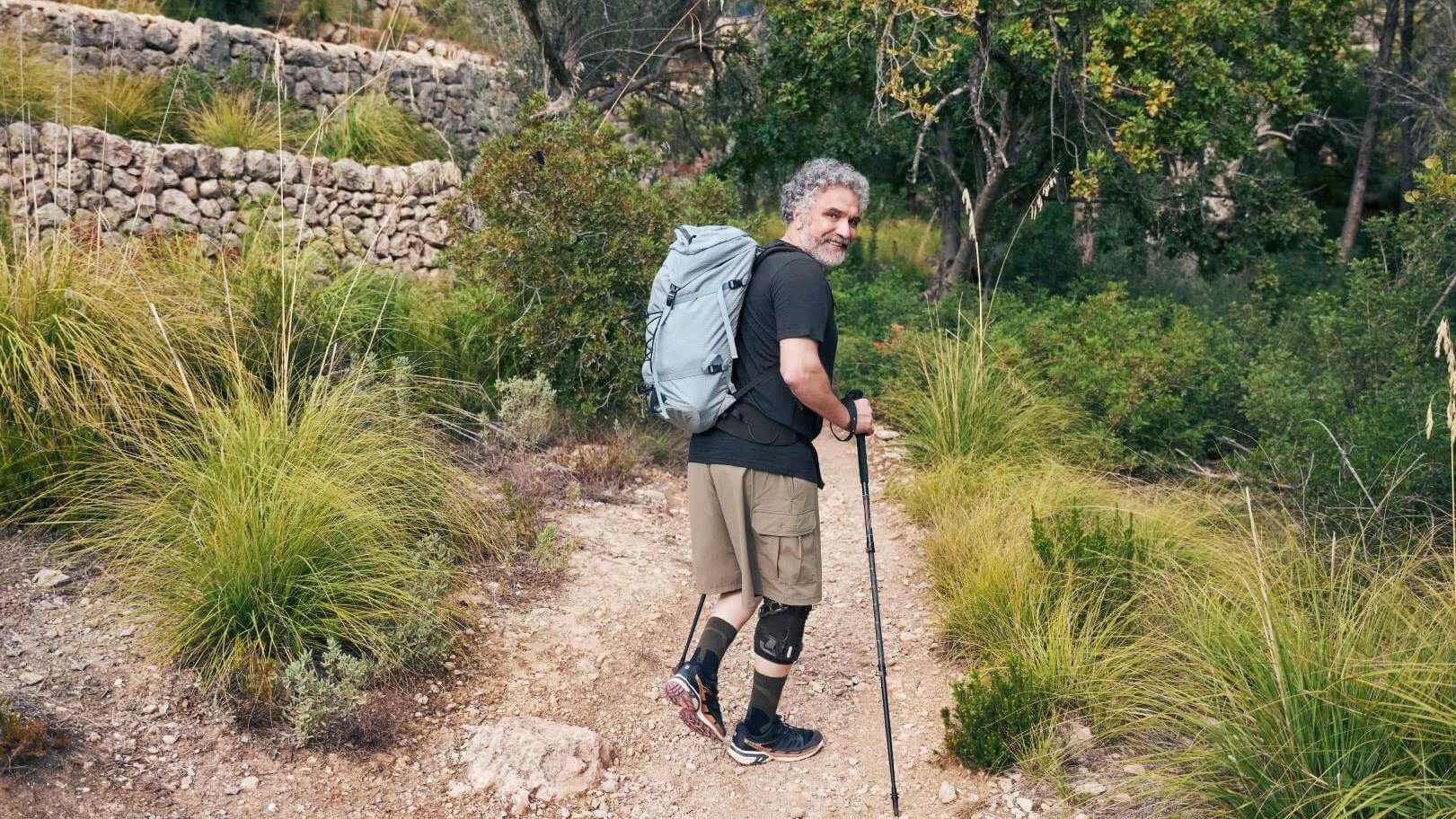 Een oudere man met grijze krullen wandelt met een trekkingrugzak en wandelstokken over een smal natuurpad door een groen heuvellandschap. Hij draagt een GenuTrain OA knieorthese ter ondersteuning bij gonartrose.