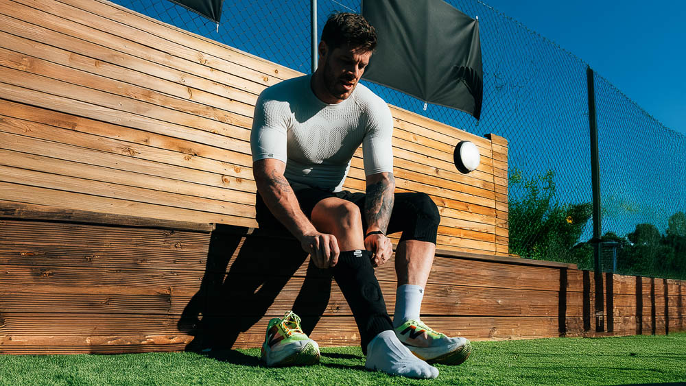 Atleet zit buiten op een houten bank en trekt een zwarte sok of compressielaag over zijn onderbeen aan; hij draagt een strak wit sportshirt en bereidt zich in de zon voor op zijn training.