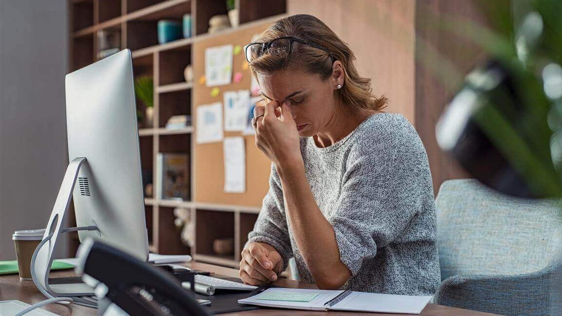 De afbeelding toont een gestreste vrouw op haar werkplek op kantoor. Ze legt haar hand tegen haar voorhoofd. Te weinig beweging, bijvoorbeeld op het werk, kan ook invloed hebben op de psyche.